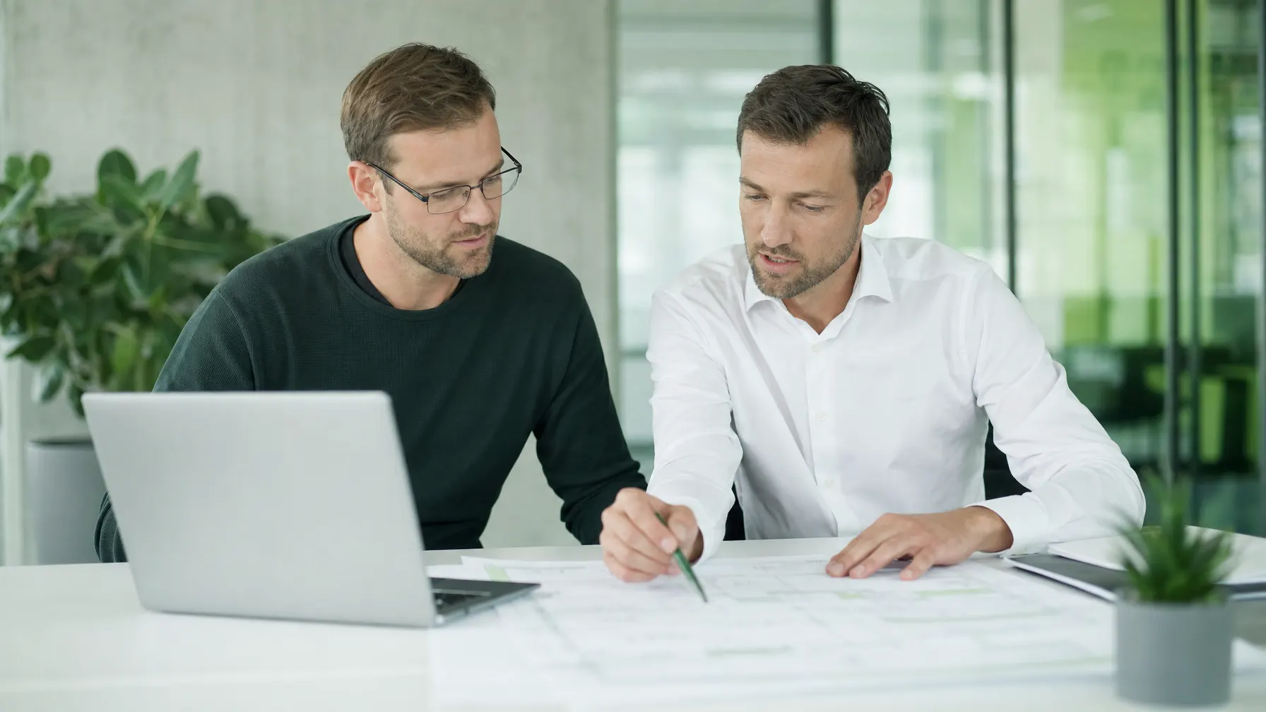IT-Berater und Kunde besprechen eine Architekturskizze am Tisch mit Laptop und Unterlagen